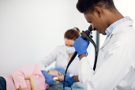Close up portrait of 30-aged Afro-American man doctor looking in endoscope camera, doing endoscopy of female patient together with nurse.の写真素材
