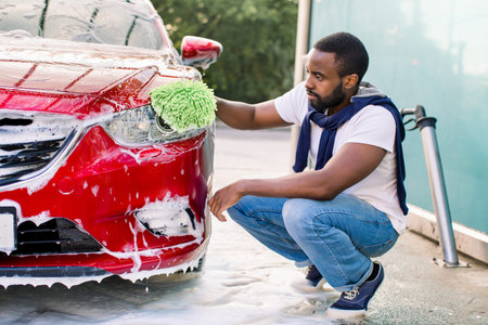 Car washing outdoors ar self wash service station. Handsome African dark skinned man in casual wear, washing his red care headlights with green sponge and soap.の写真素材