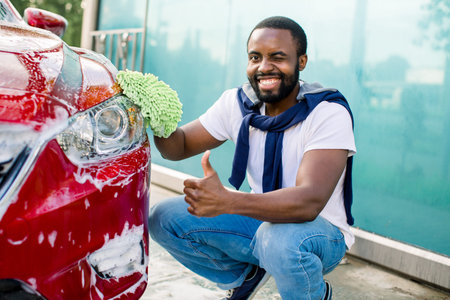 Young cheerful African man posing to camera with thumb up, while cleaning his luxury red car outdoors at self car wash station, using green rag and soap foam.の写真素材