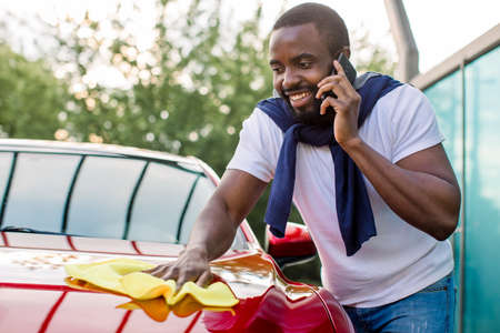 Washing car outdoor, self service station. Young busy African man talking phone while polishing his red car with yellow microfiber clothの写真素材