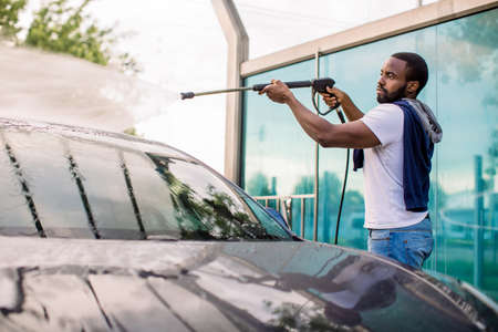 Handsome African young man in casual wear, washing his red car manually with water high-pressure hose at outdoor self wash service. Car washing concept.の写真素材
