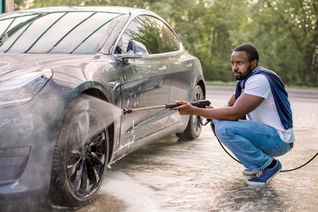 Young African man in casual wear washing his luxury modern electric car at self car wash station outdoors, using high pressure water spray.の写真素材