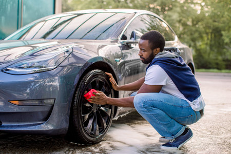 Car wash service outdoors. Car wash self-service concept. Handsome African guy wiping wheel of his...
