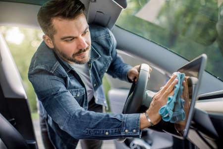 Young handsome Caucasian man cleaning the car console and touchscreen display of his modern electric car with microfiber cloth. Car wash and detailing concept at self car wash station.の写真素材