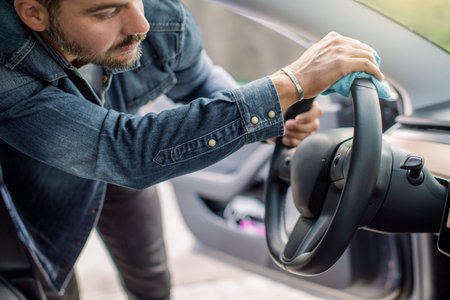 Young handsome bearded man, cleaning his modern futuristic car interior, car steering wheel with...