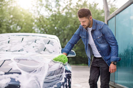 Outdoor car wash concept. Portrait of young man in casual wear, washing his luxury modern car in a self-service car wash station with sponge and foamの写真素材