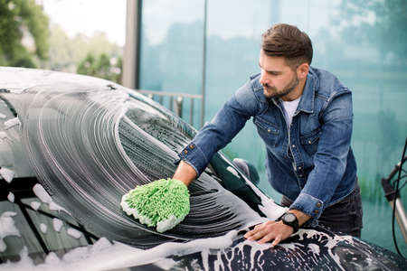 Handsome young Caucasian man cleaning his car windshield with green sponge mitten and soap foam outdoors at car wash service.の写真素材