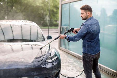 Handsome guy in jeans shirt washing his modern electric luxury car, rinsing the soap with high pressure water jet. Manual car wash with pressurized water in car wash outsideの写真素材