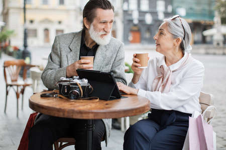 Stylish aged couple relaxing together on cafe terrace and enjoying hot coffee. Lovely wife and husband using digital tablet and retro camera outdoors.の写真素材