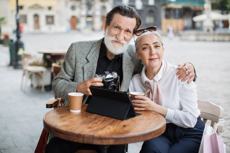 Pleasant senior couple hugging and smiling while sitting at cafe table with digital tablet and retro...