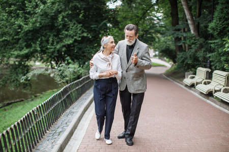 Full length portrait of happy senior couple walking and talking at green park. Bearded husband gently hugging beautiful wife. Pensioners enjoying time together.の写真素材