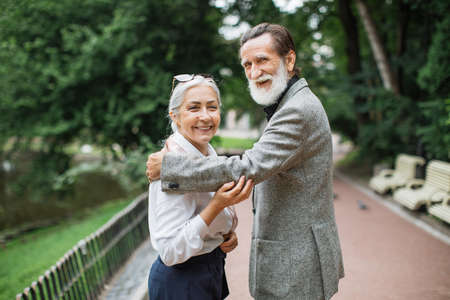 Charming aged husband and wife smiling and looking at camera while standing at green park. Concept of relationship, retirement and togetherness.の写真素材