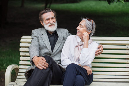 Romantic aged couple sitting on wooden bench at green park and talking. Mature family enjoying time together. Happy retirement.の写真素材
