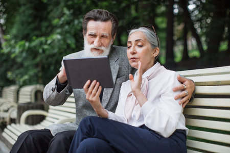 Beautiful caucasian family sitting on bench, looking on each other and holding digital tablet. Elder...