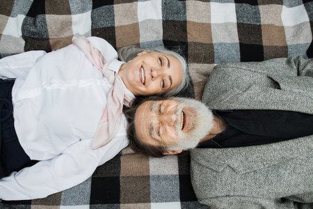 Top view of caucasian mature couple lying head to head on checkered blanket and smiling on camera....