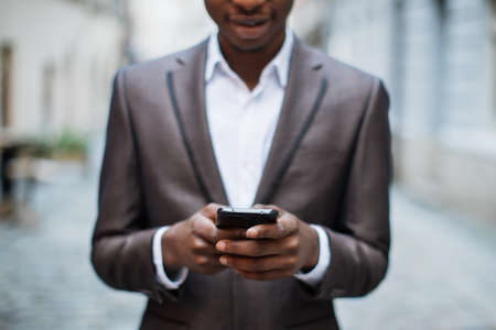 Close up of happy african man dressed in elegant suit holding modern smartphone in hands while standing outdoors. Concept of people, technology and business.の写真素材