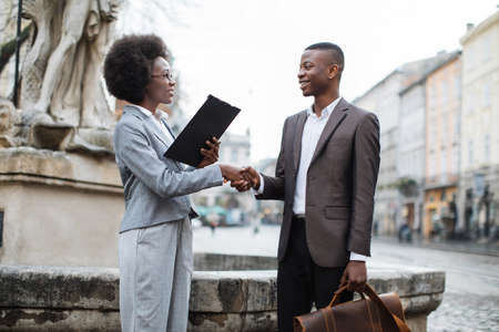 Successful businesswoman and businessman in stylish formal clothes shaking hands on street. Two african partners finishing successful meeting outdoors.の写真素材