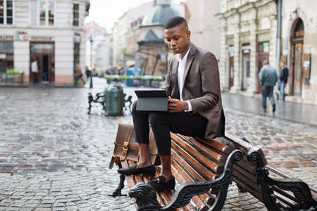 Confident african businessman using digital tablet for remote work while sitting on wooden bench outdoors. Concept of modern technology.の写真素材