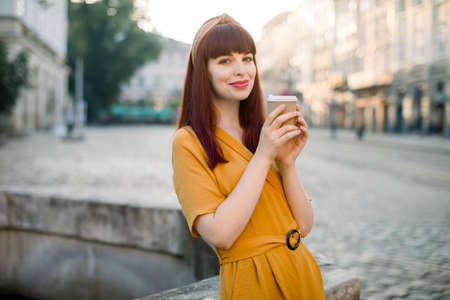 Outdoor portrait of Caucasian woman in yellow dress, with takeaway coffee cup at the city. Smiling pretty dark haired girl posing to camera in old city, enjoying a cup of coffee, hot chocolate.の写真素材