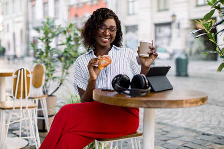 Pretty African business lady having lunch time outdoor in city cafe. African American smiling woman sitting at the table in city cafe, holding a cup of coffee and croissant, looking at camera.の写真素材