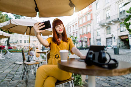 Charming young pretty woman in yellow overalls, making selfie photo or having video call, while sitting in outdoor city cafe, drinking coffee and using digital tablet.の写真素材