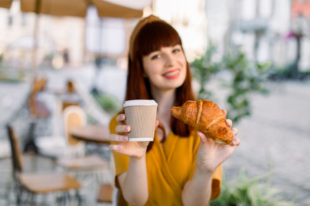 Outdoor city lifestyle portrait of young attractive Caucasian businesswoman, sitting at cafe with paper takeaway cup of coffee and fresh croissant, smiling to camera. Focus on the cup and croissant.の写真素材