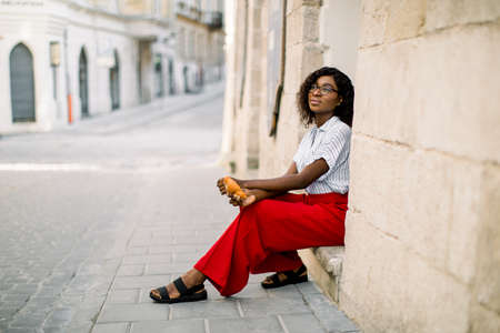 Horizontal shot of young attractive dreamy African woman dressed in red pants and light shirt, sitting on the stairs of old vintage building on the street outdoor, holding croissant.の写真素材