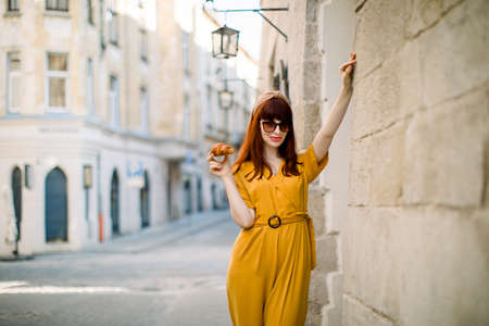 Fashion city portrait of gorgeous young Caucasian red haired woman, dressed in stylish yellow overalls, sunglasses and head hoop, walking in old European city with fresh tasty croissant.の写真素材