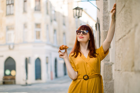 Outdoor city portrait of dreamy pretty young Caucasian red haired woman, dressed in stylish yellow overalls, sunglasses and head hoop, walking in old European city with fresh tasty croissant.の写真素材