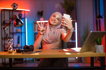 Happy arabian woman in headscarf gesturing sign peace while taking selfie modern smartphone. Young...