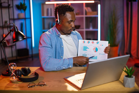Good-looking confident black-skinned businessman in wireless headset, sitting at the desk with...
