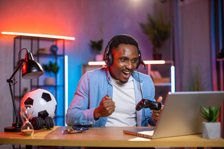 Handsome excited joyful young African man in headset celebrating victory in video game on laptop with clenched fists and smile, spending his leisure at home in the eveningの写真素材