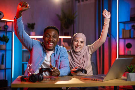 Joyful black man and arabian woman feeling happiness while winning in video games on laptop. Multi ethnic couple enjoying entertainment at home during evening time.の写真素材