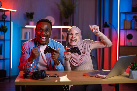Joyful African man and Muslim woman feeling happiness while winning in video games on laptop. Multi...