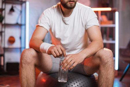 Cropped close shot of handsome sporty man wearing white t-shirt and gray trousers, sitting on fitball and holding in hands bottle with mineral water. Refreshing and sport conceptの写真素材