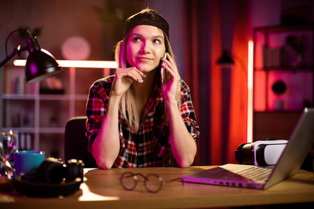 Attractive caucasian woman talking on modern smartphone while sitting at desk with wireless laptop....