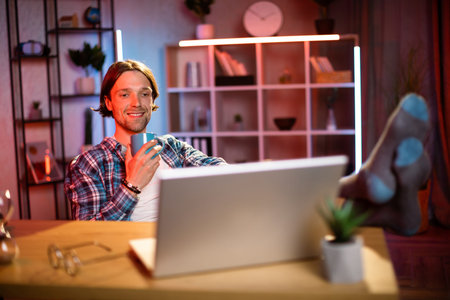 Attractive young man sitting at desk with mug of hot drink in hand and looking at laptop screen....
