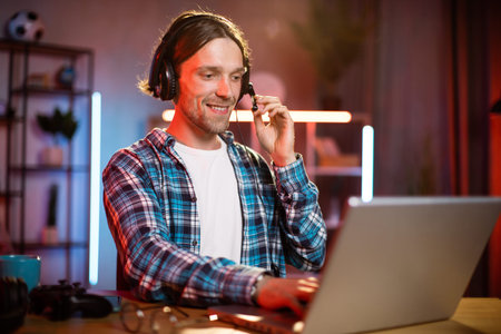 Portrait of young caucasian man in headset sitting at desk and using modern laptop for remote work at home. Male freelancer looking at laptop screen with serious facial expression.の写真素材