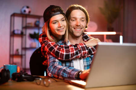 Focused caucasian man working on modern laptop during evening time and his girlfriend hugs him from back. Concept of people, home routine, freelance and deadline.の写真素材