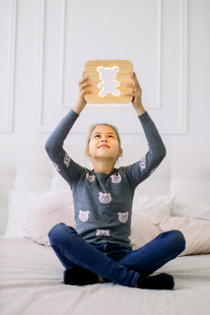 Indoor portrait of pretty school girl in casual domestic wear, sitting on the bed in lotus position and holding stylish wooden night lamp with bear picture under her headの写真素材