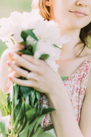 Beautiful young Caucasian blond woman in light dress holding bouquet of white peonies, walking in summer field or garden in sunset. Woman with flowers outdoors.の写真素材