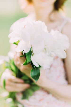 Beautiful young Caucasian blond woman in light dress holding bouquet of white peonies, walking in summer field or garden in sunset. Woman with flowers outdoors.の写真素材