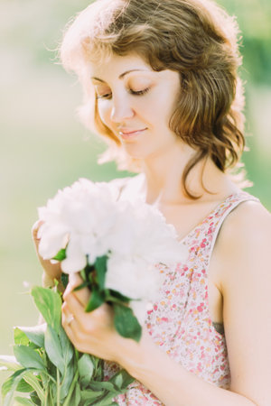 Beautiful young Caucasian blond woman in light dress holding bouquet of white peonies, walking in summer field or garden in sunset. Woman with flowers outdoors.の写真素材