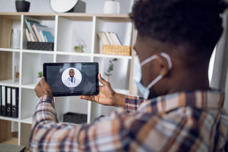 View from shoulder of african man in face mask sitting on couch and using digital tablet for calling...