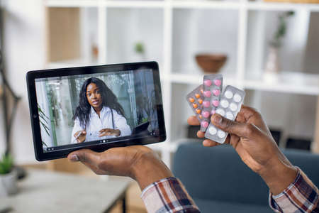 Close up of male hands holding various pills and digital tablet with afro american doctor on screen. Sick person calling therapist for online consultation about health treatment.の写真素材