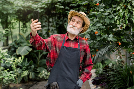 Portrait of handsome cheerful 60s bearded man gardener, wearing checkered shirt, apron and straw...