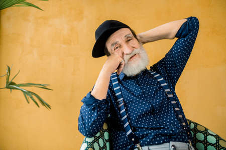 Fashion studio portrait of happy stylish 70-aged man in black cap, sitting on colorful chair on yellow background, posing on camera with pleasant smile and arms behind his head.の写真素材