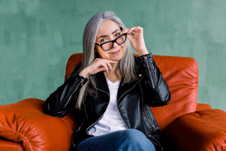Pretty smiling 60-aged senior lady with long straight gray hair, looking at camera, while sitting in...