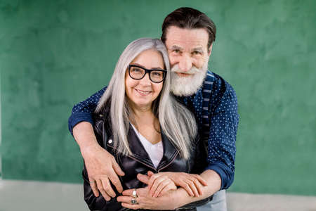 Studio shot of cheerful stylish bearded man embracing his gray haired charming wife from the back and looking at camera with smile, standing on grenn wall background.の写真素材