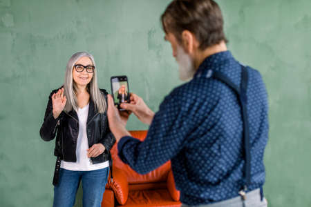 Handsome bearded senior man making photo on the phone for his beautiful wife with long gray hair, posing on the background of green wall near red chair and waving to camera.の写真素材